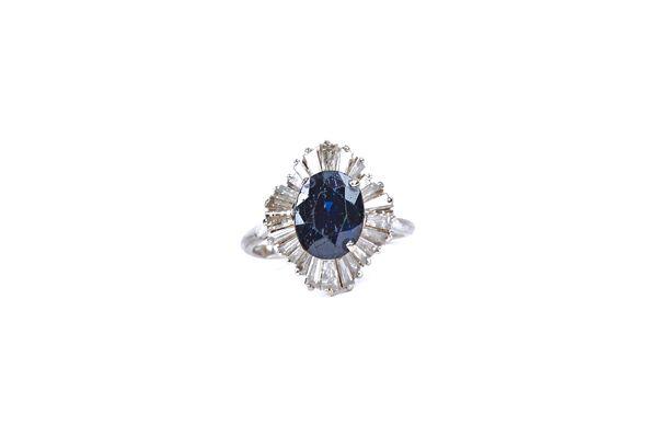 A white gold, sapphire and diamond ring, claw set with the oval cut sapphire at the centre in a radiating surround, mounted with baguette diamonds, de