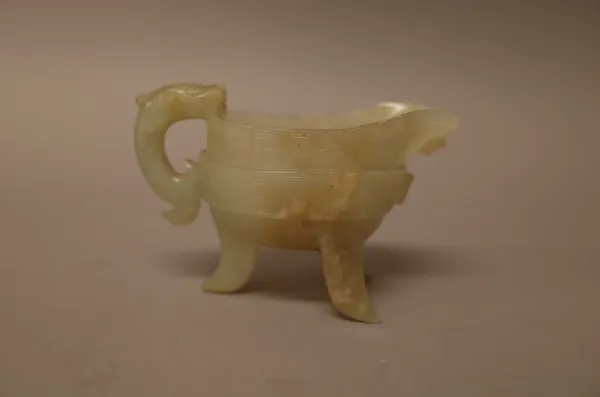 A Chinese archaistic jade libation cup, raised on three splayed supports, the sides carved with a band of taotie masks, the stone of pale celadon tone