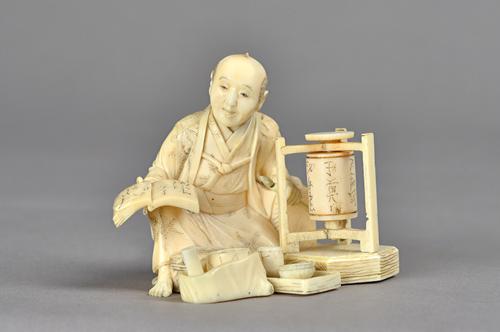 A Japanese ivory okimono of a seated calligrapher, Meiji period, seated beside a revolving cylinder decorated with characters, holding a brush and ope