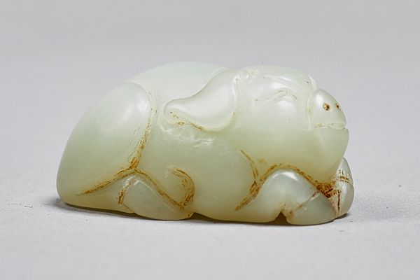 A Chinese celadon jade figure of a pig, Qing Dynasty, carved in recumbent pose with front trotters tucked under its chin, 5cm. length. Illustrated. Pr