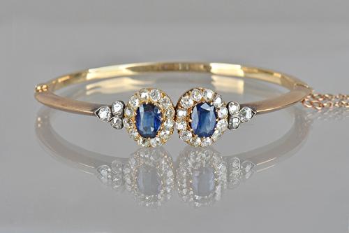 A gold, sapphire and diamond set oval hinged bangle, in a twin cluster design, each cluster claw set with a cushion shaped sapphire to the centre, in