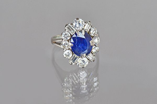 A sapphire and diamond oval cluster ring, claw set with the oval cut sapphire in a surround of eight circular cut diamonds and with four baguette diam