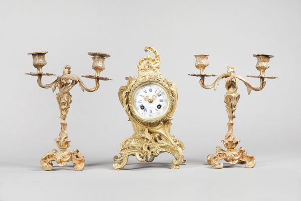 A French gilt brass clock garnitureIn the Louis XV style, circa 1875The clock of shaped outline with  3 1/2 in. circular white enamel dial with blue R