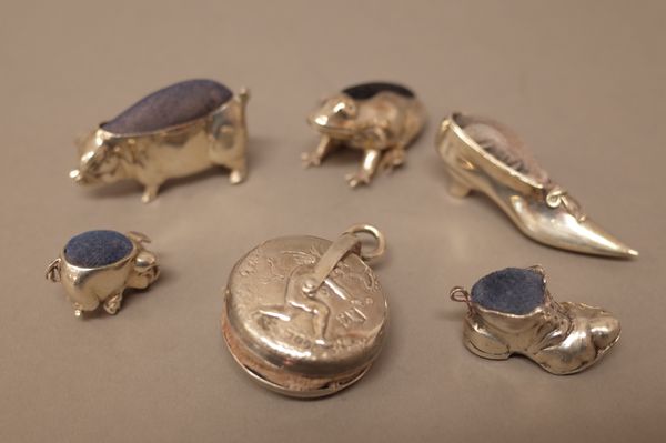 A silver mounted circular pin cushion, decorated with cherubs, import mark London 1900, four silver mounted modern pin cushions, comprising; a pig, a