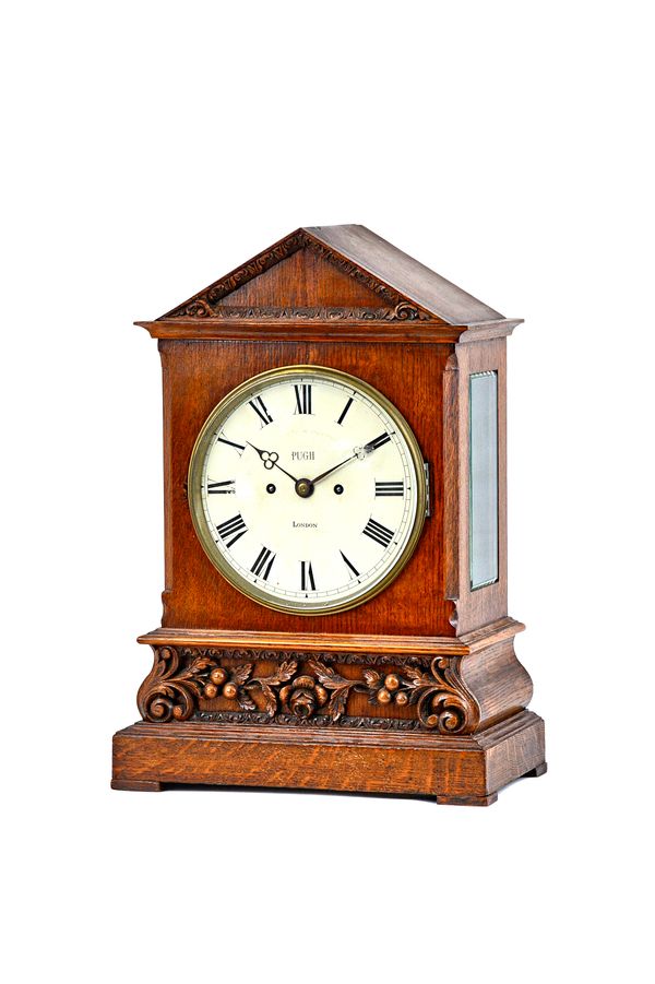 A German oak cased mantel clock with painted metal dial detailed 'Pugh London' over a frieze ornately carved with fruiting vines, enclosing a twin fus