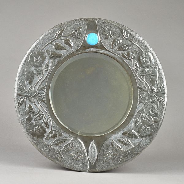 A Liberty style Arts and Crafts pewter wall mirror, circa. 1900, of circular form with bevelled glass relief embossed foliate border and inset with a