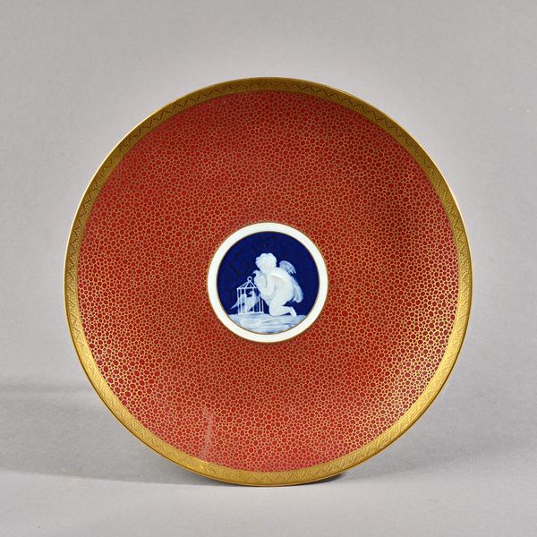 A Mintons pâte-sur-pâte plate, circa 1884, decorated in the centre with a deep blue ground circular panel depicting a kneeling cupid with two birds an