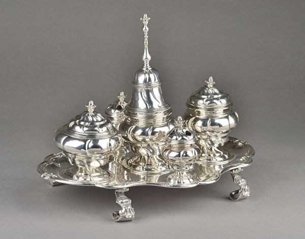 A Spanish inkstand, of shaped oval form, having a bold scrolling border, raised on four bold Rococo feet, the central sander with a bell form cover, t