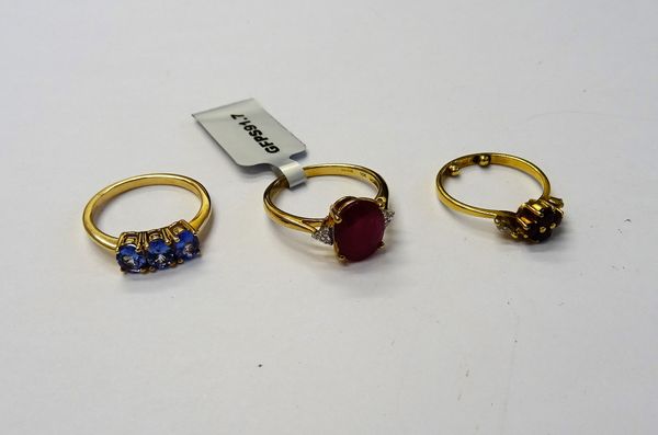 A 9ct gold ring, claw set with a row of three oval cut tanzanites, a 9ct gold ring, claw set with an oval cut treated ruby, between colourless gem set