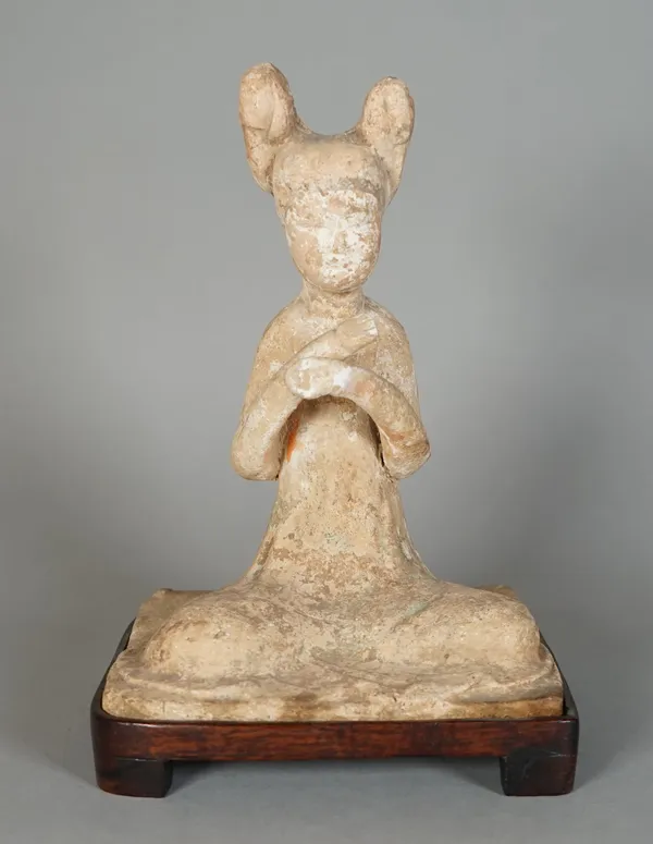 A Chinese pottery figure of a maiden, probably Tang dynasty, modelled seated with hands crossed, her hair bound up in two buns, 17.5cm. high, wood sta