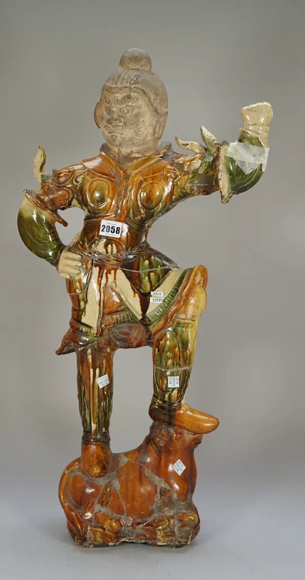 A large Chinese sancai glazed pottery tomb guardian, possibly Tang dynasty, with unglazed head, standing in body armour on a recumbent ox,  his right