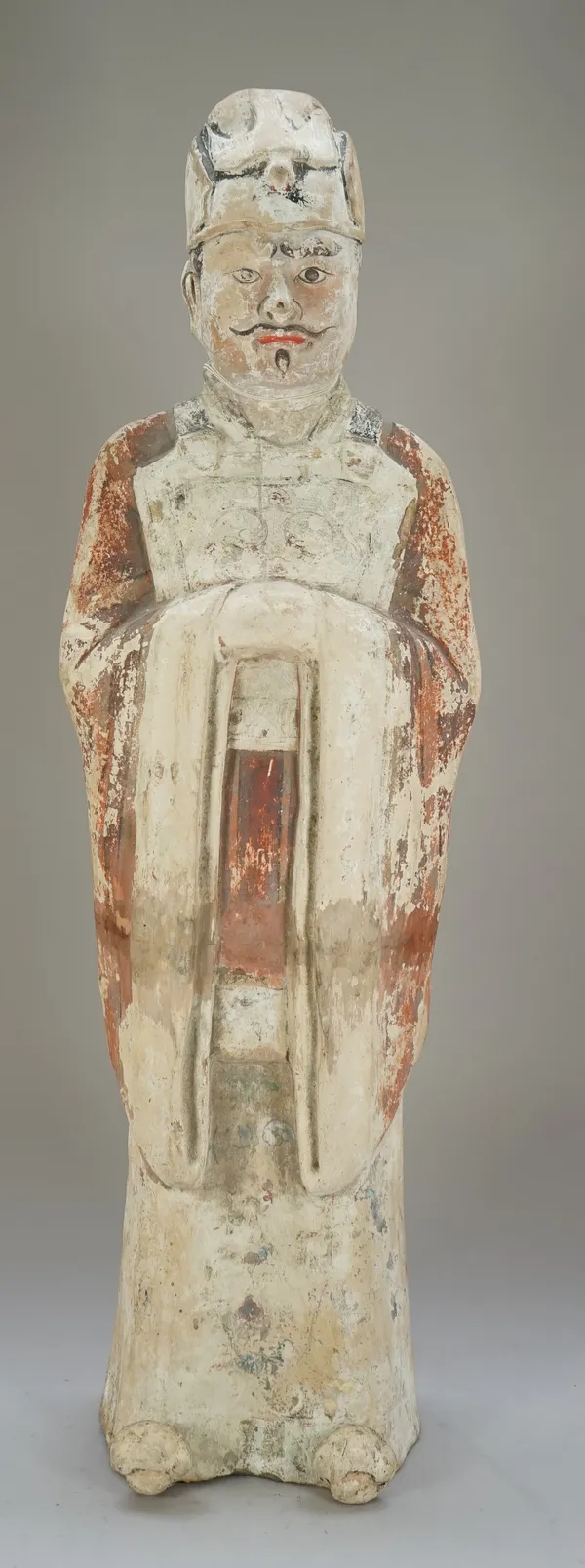 A tall Chinese painted pottery official, probably Tang dynasty, standing in long robes with hands clasped, wearing a cap with bird motif, painted red