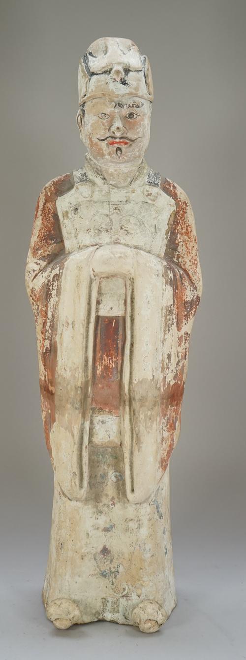 A tall Chinese painted pottery official, probably Tang dynasty, standing in long robes with hands clasped, wearing a cap with bird motif, painted red