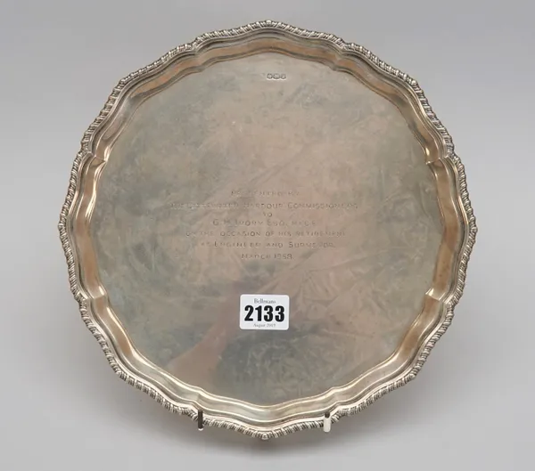 A silver salver, of shaped circular form, having a shaped pie-crust rim with a gadrooned border, presentation inscribed to the centre, raised on four