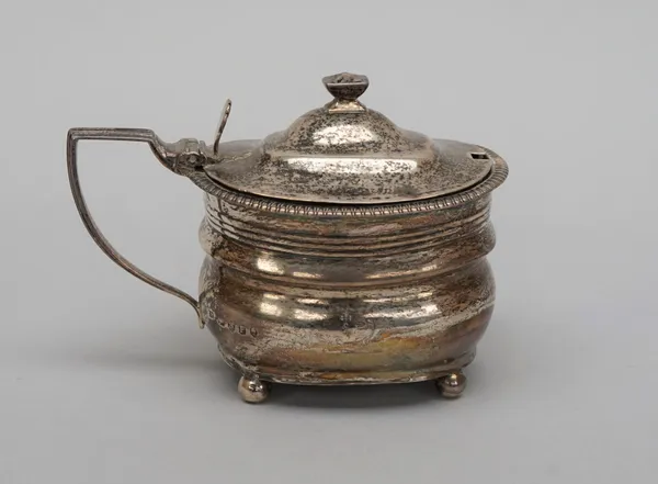 A George III mustard pot, of curved form, decorated with a reeded band, with a shell thumbpiece and raised on four spherical feet, crest engraved, Lon