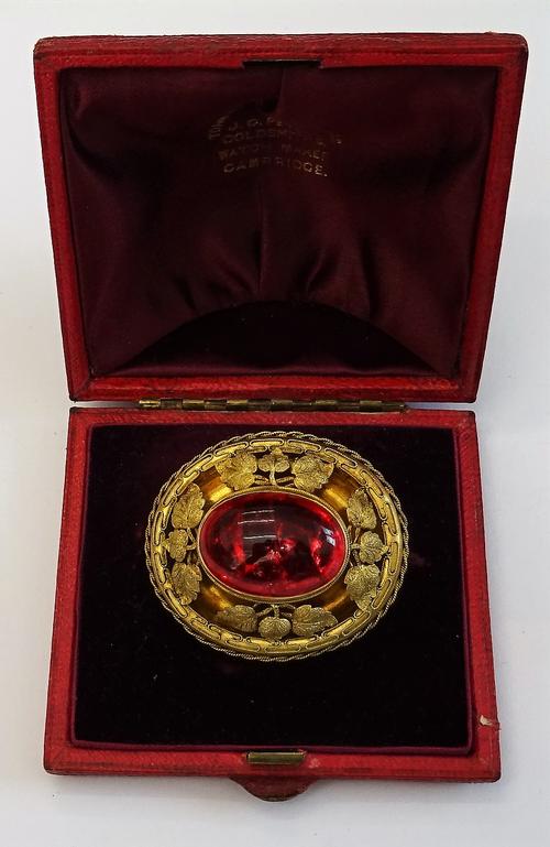 A Victorian gilt and cabochon stained red gem set solitaire brooch, of oval form, decorated with four foliate sprays at intervals, within ropetwist ap