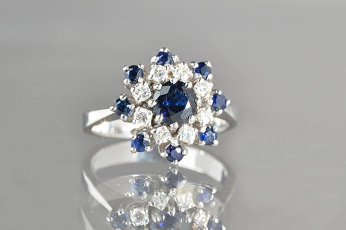 A white gold, sapphire and diamond set shaped circular cluster ring, claw set with the principal circular cut sapphire to the centre, in a surround of