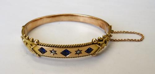 A 9ct gold, sapphire and diamond set oval hinged bangle, mounted with three cushion shaped sapphires and with two rose diamonds at intervals, otherwis