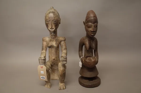 An African tribal female figure modelled seated and holding an umbrella, 51cm high and one further female carved wood figure holding a bowl, 48cm high