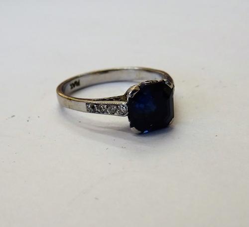 A platinum, sapphire and diamond ring, claw set with the cut cornered square step cut sapphire at the centre, between diamond set four stone shoulders