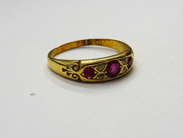 A gold, ruby and diamond ring, mounted with three cushion shaped rubies alternating with two pairs of smaller cushion shaped diamonds, between scrolle