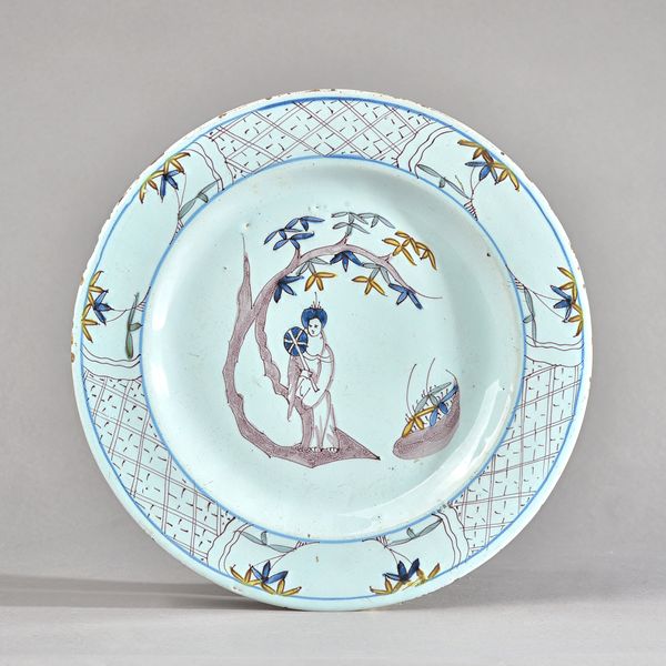 A Bristol delftware polychrome plate, circa 1760, painted in the centre with a woman with a fan standing against a tree, inside a diaper pattern borde