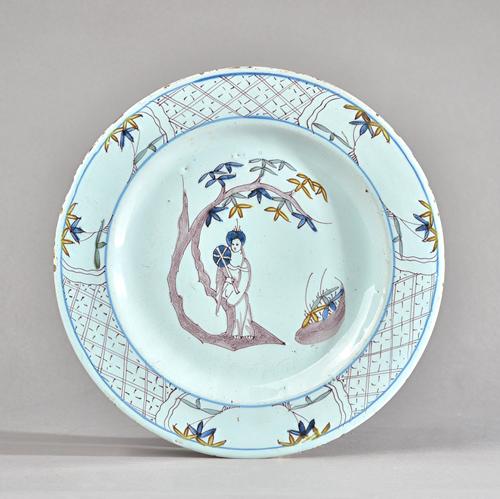 A Bristol delftware polychrome plate, circa 1760, painted in the centre with a woman with a fan standing against a tree, inside a diaper pattern borde