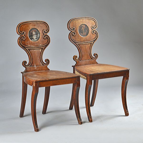 A pair of George III mahogany hall chairs, with shaped backs inset with rubbed painted heraldic crests, on sabre supports,  46cm wide x 88cm high, (2)