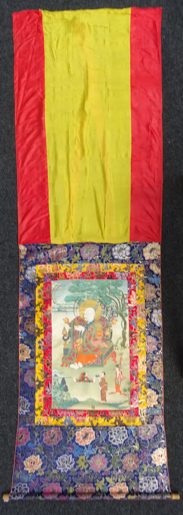 A Tibetan thangka depicting an arhat, 20th century, pigment with gold on cloth, the elderly figure seated on an elaborate throne under a flowering tre