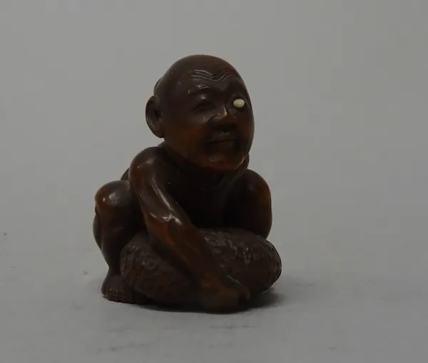 A Japanese wood netsuke of a blind beggar, 19th century, attempting to lift his distended scrotum, one eye with ivory inlay, signed Gyokkei on ivory t