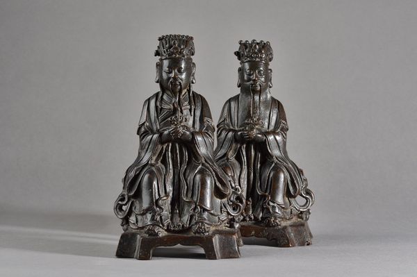 A pair of Chinese bronze figures of officials, Ming dynasty, 17th century, each bearded figure seated on a bench with hands clasped, raised on a brack