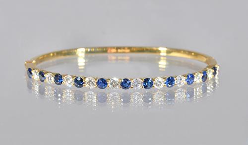 An 18ct gold, sapphire and diamond oval hinged bangle, claw set with a row of circular cut diamonds alternating with circular cut sapphires, on a snap