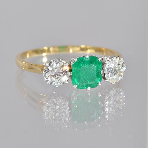 A gold, emerald and diamond set three stone ring, claw set with the cut cornered square step cut emerald to the centre, between two claw set cushion s