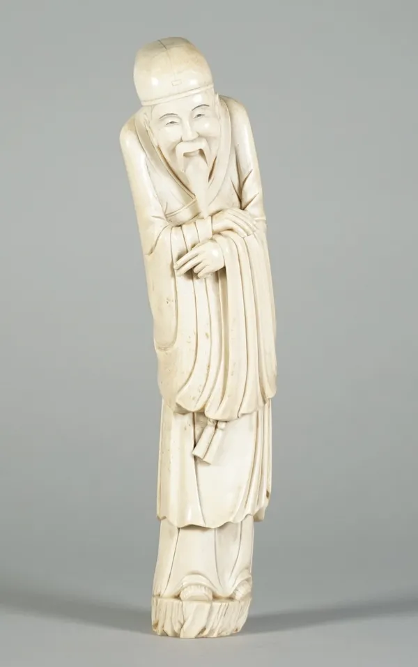 A Chinese ivory figure of an Immortal, 19th century carved wearing long robes and a cloth hat, his arms folded, 27.6cm. high. Illustrated 10