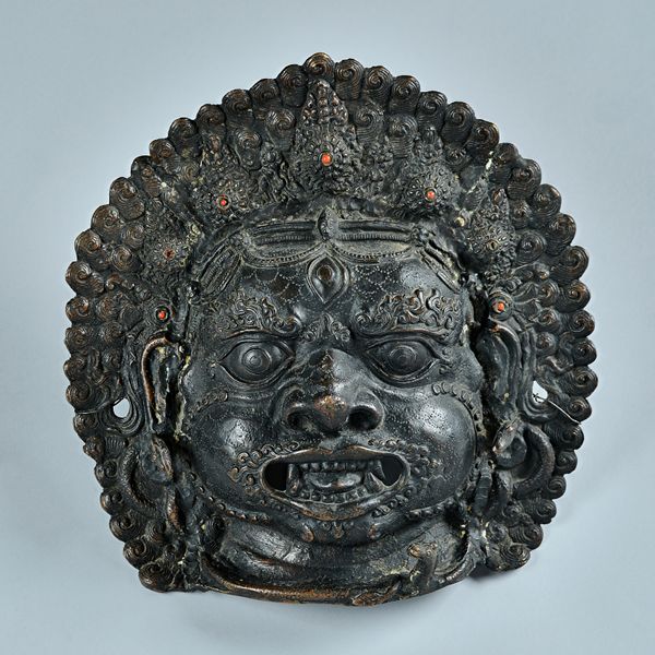 A Nepalese bronze mask of Bhairava, 19th century, the Hindu deity with flaming hair inset with orange `jewels', bulging eyes, bared teeth and with sna