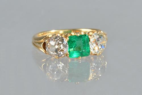 A gold, emerald and diamond set three stone ring, claw set with a square step cut emerald to the centre, between two cushion shaped diamonds, between