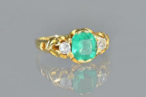 A gold, emerald and diamond set three stone ring, mounted with the step cut emerald at the centre, between two cushion shaped diamonds, between scroll