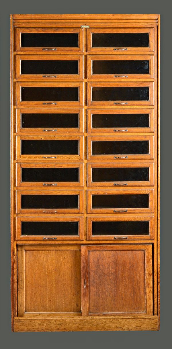 Dudley & Co Ltd, A mid-20th century oak haberdashery cabinet with two rows of eight glass fronted drawers over sliding cupboards, 91cm wide x 193cm hi