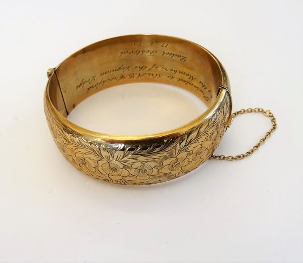 A 9ct gold hollow bangle of oval form, engraved to one side with repeat flowerheads and foliage, the inner band presentation inscribed, fastened with