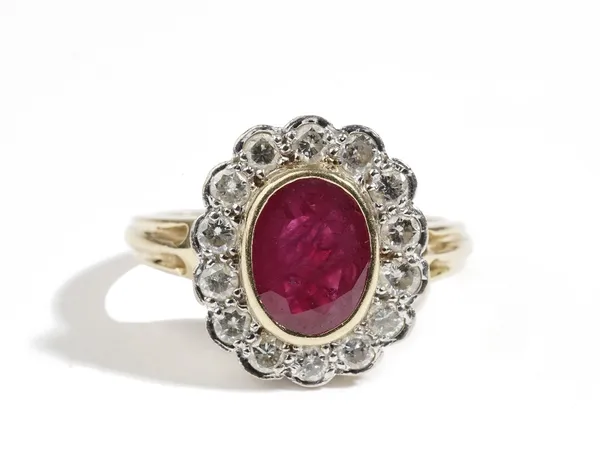 An 18ct gold, ruby and diamond oval cluster ring, collet set with the oval cut ruby in a surround of circular cut diamonds, between ridged shoulders,