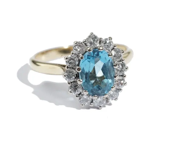 An 18 ct gold, blue topaz and diamond oval cluster ring, claw set with the oval cut blue topaz in a surround of twelve circular cut diamonds, London 1