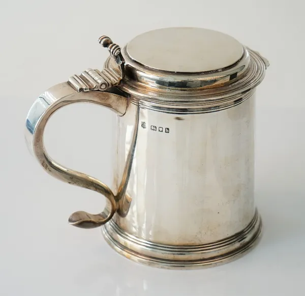 A silver hinge lidded tankard of tapered cylindrical form with a cast scrolling thumb piece to the scrolled handle, with a flat lid and with a circula