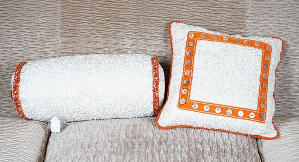 A scrunched cream silk bolster cushion, with braided ends, and another of square shape with a border of mother of pearl buttons (2).