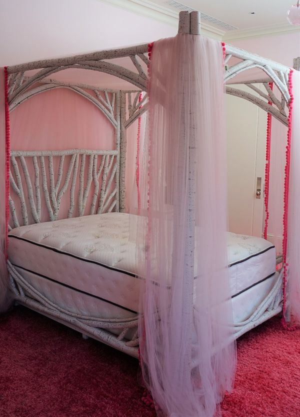 A four poster bed, the faux white birch rustic frame supporting a canopy and hung with white net curtains trimmed with pink bobbles, with a Beautyrest