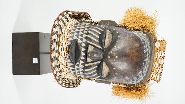 Tribal interest; an African tribal wooden mask decorated all over with cowrie shells, straw and brass studs to the brow and nose, on a wooden stand, m