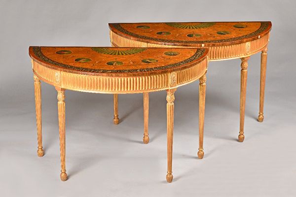 A pair of George III style demi-lune console tables, each with polychrome painted satinwood top with ebonised border, the gilt base with fluted frieze