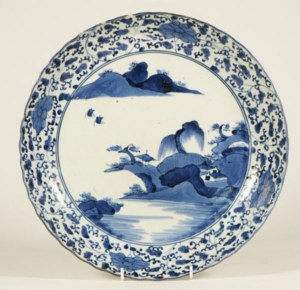 An Arita blue and white dish, Edo period, painted with a river landscape beneath a border filled with flowers and tendrils, (a.f), 31.5cm. diameter.