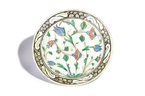 An Iznik pottery dish, early 17th century, painted in blue, green, red and black with flowers issuing from a leafy tuft inside a breaking wave border,