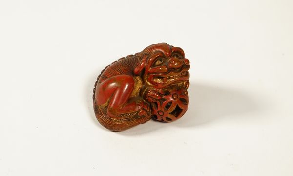 A Japanese red lacquered wood  netsuke of a shishi, 19th/early 20th century, carved in recumbent pose with front paws resting on a brocade ball, gilt