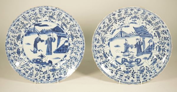 A pair of Chinese blue and white dishes, Kangxi, each painted in the centre with a man an woman in a garden beside a pavilion, beneath a moulded borde